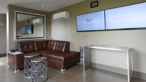 Un área de espera moderna con un sofá de cuero marrón, una mesa de centro de metal y una consola blanca. Un televisor de pantalla plana en la pared muestra el texto "JUSTE POUR LE FUN".