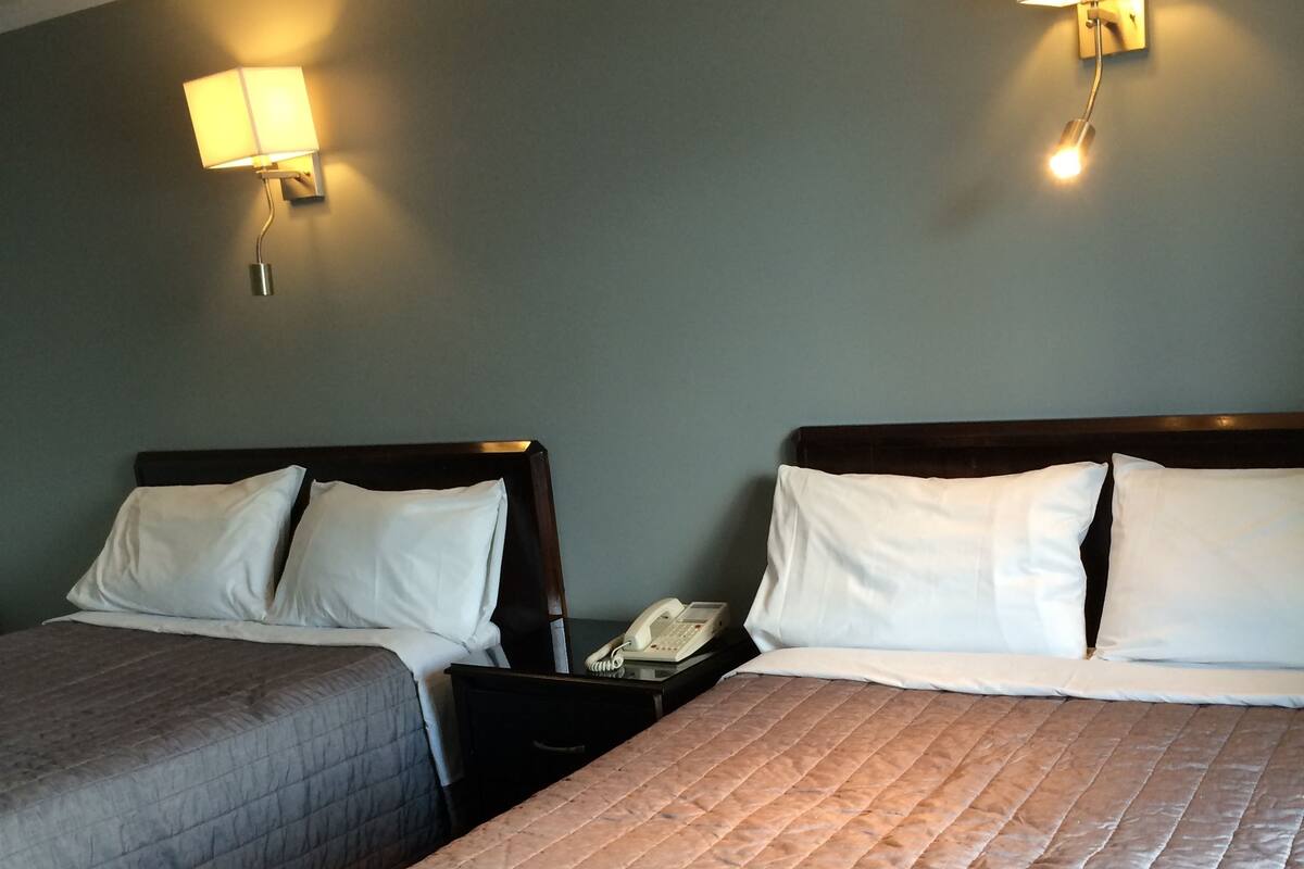 A hotel room with two beds, each with a headboard and white bedding. There are wall-mounted lamps on either side of the beds.