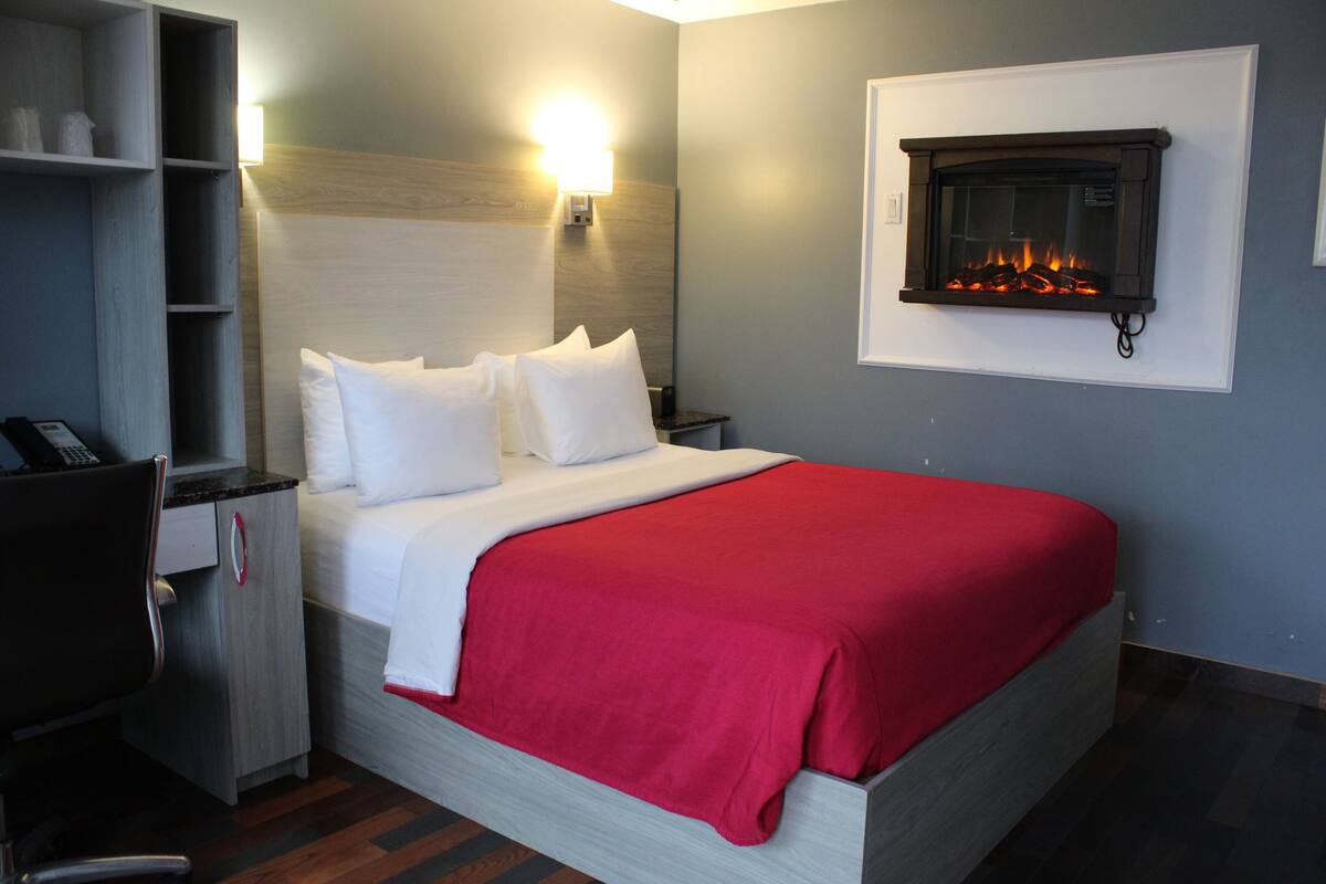 A hotel room with a bed, a desk, a chair, a nightstand, a wall-mounted fireplace, and a shelf with items.