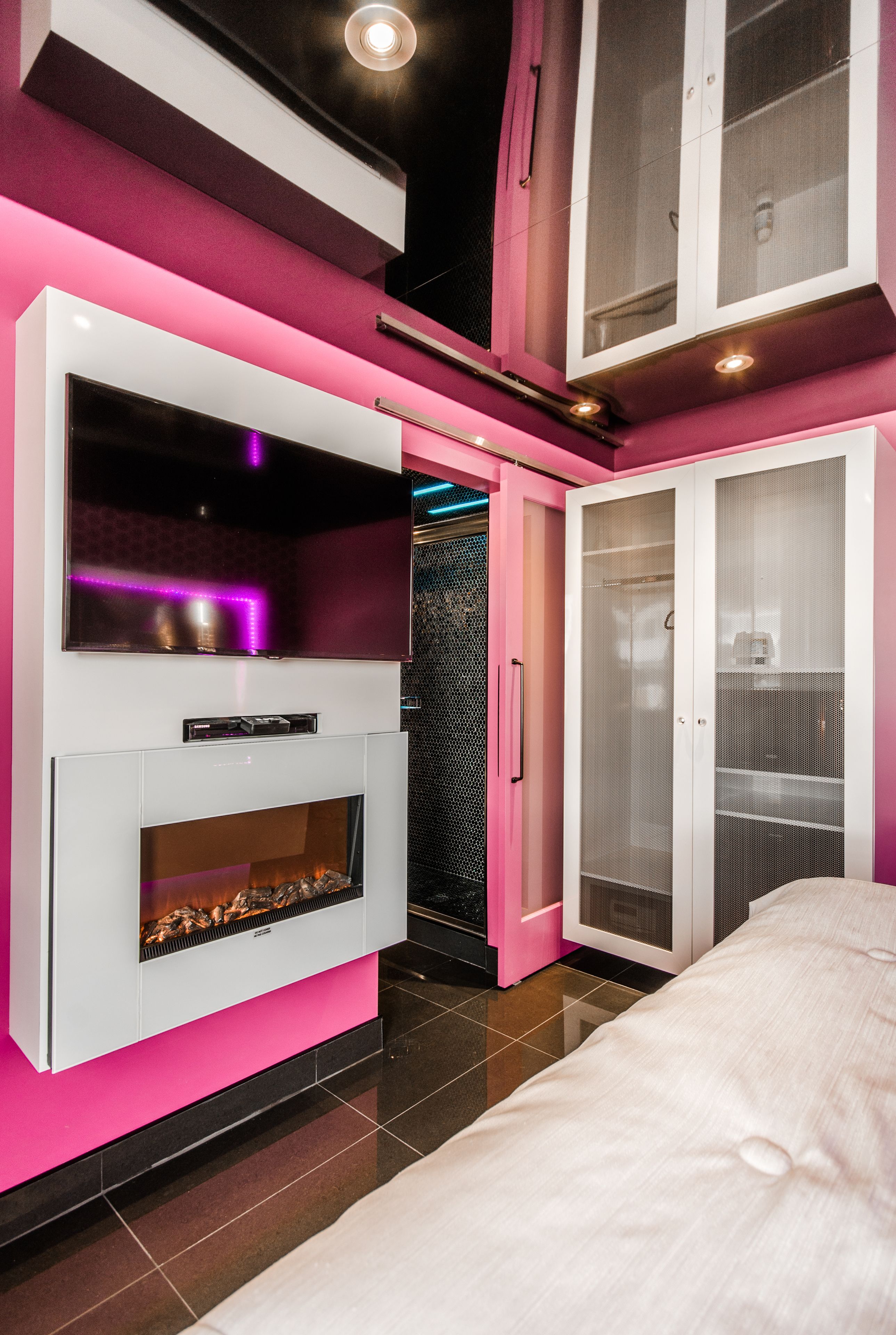 A modern bedroom with a pink and white color scheme, a built-in fireplace, a TV, and a wardrobe with a mesh door.