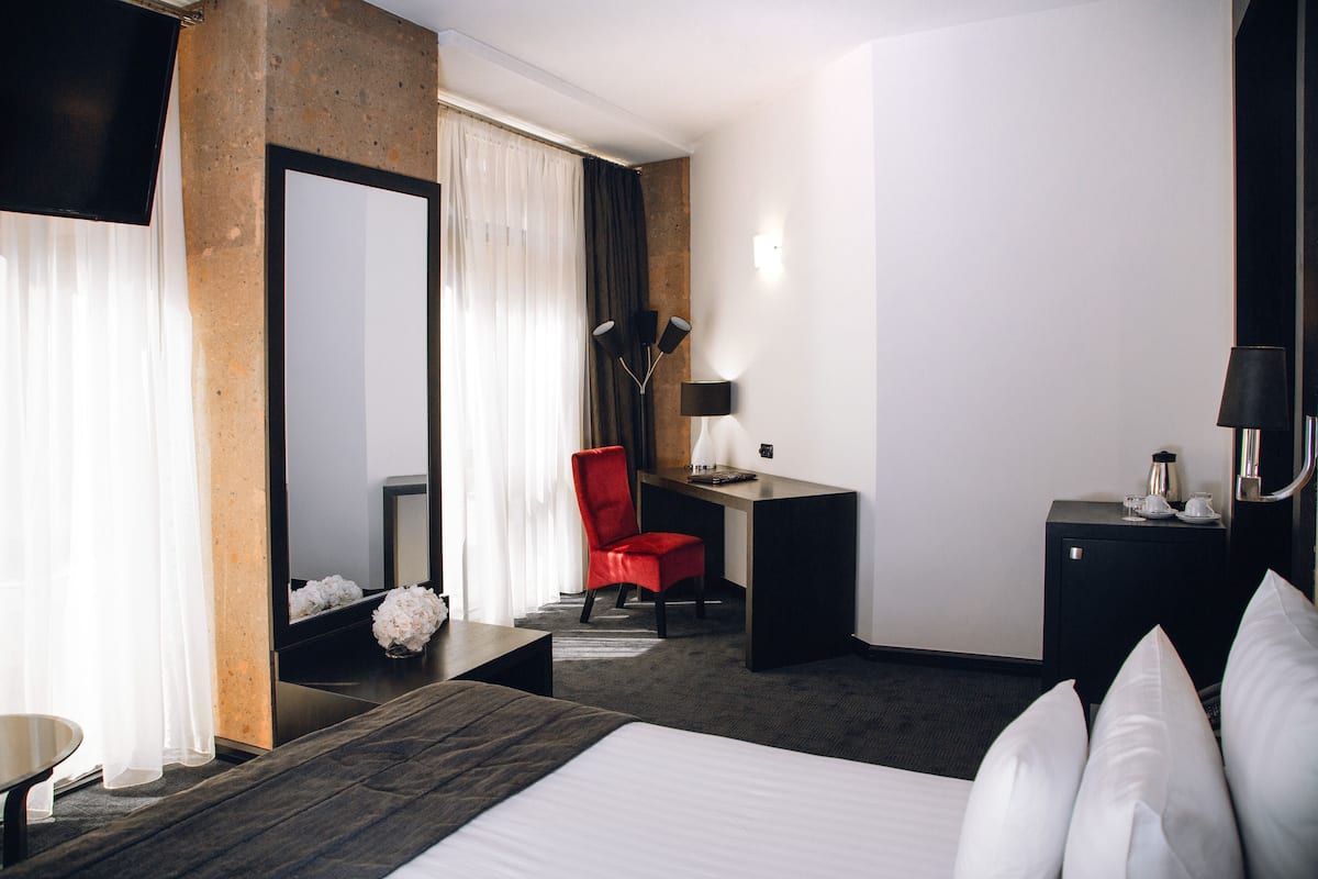 Una habitación de hotel con una cama grande, un escritorio, una silla roja, una mesita pequeña, un espejo y una mesita de noche con una lámpara.