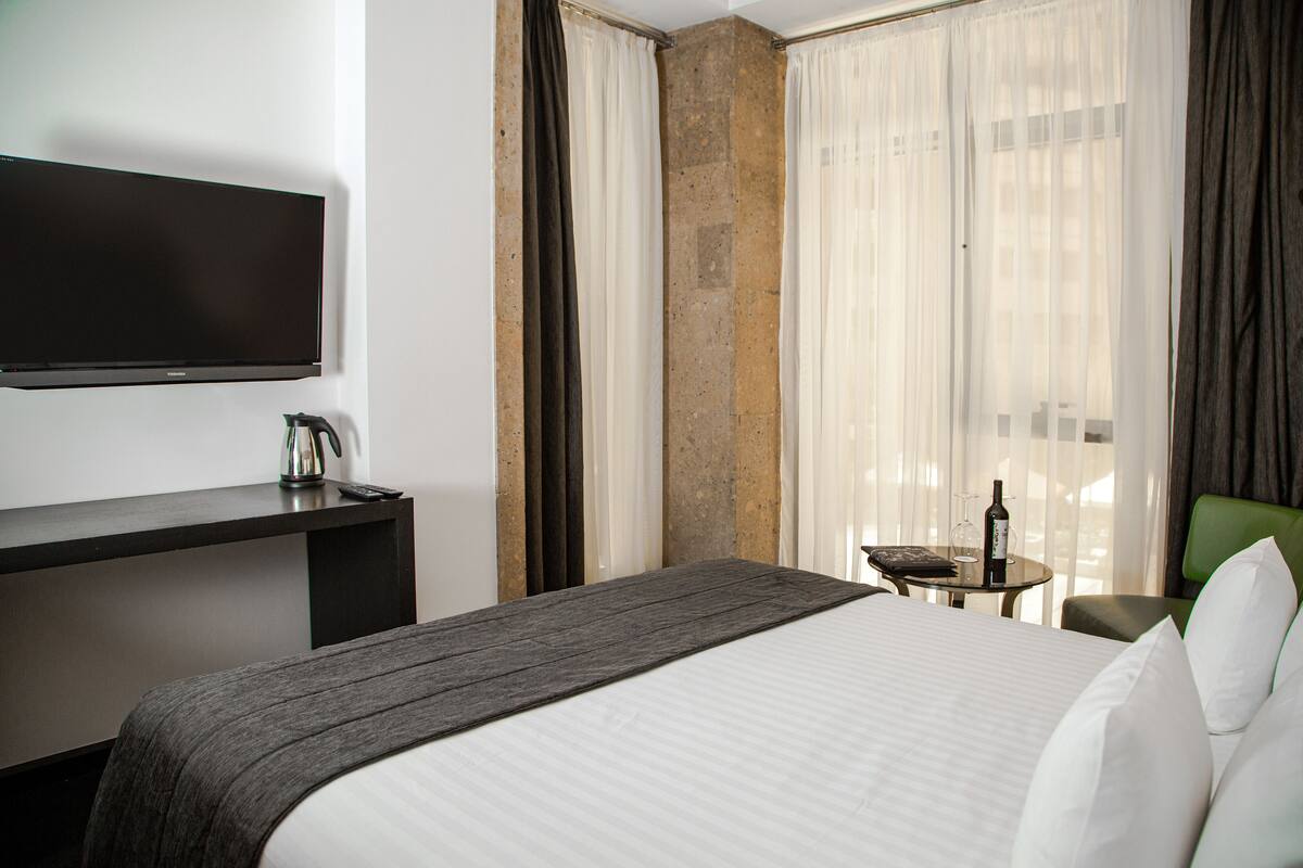 Habitación de hotel con cama, escritorio con televisor de pantalla plana, pequeña mesa con una botella y un vaso, y una silla.