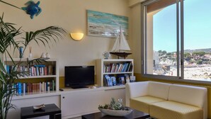A living room with a TV, bookshelves, a sofa, and a large window with a beach view. - Golf Hôtel (Bandol)