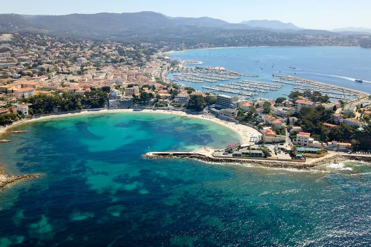 Vue aérienne d’une ville côtière avec une baie turquoise aux eaux limpides, un port de plaisance et des immeubles résidentiels.