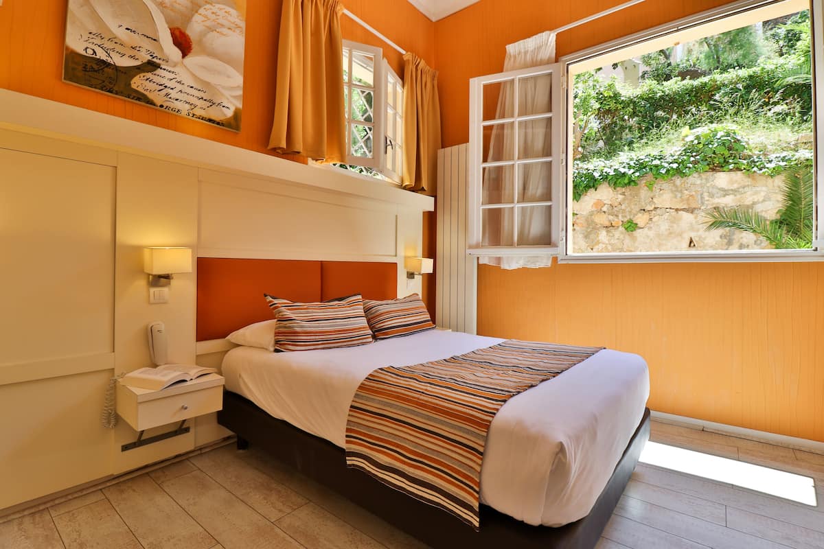 Une chambre à coucher avec un lit, une table de chevet, une fenêtre avec des rideaux et une vue sur la verdure à l’extérieur.