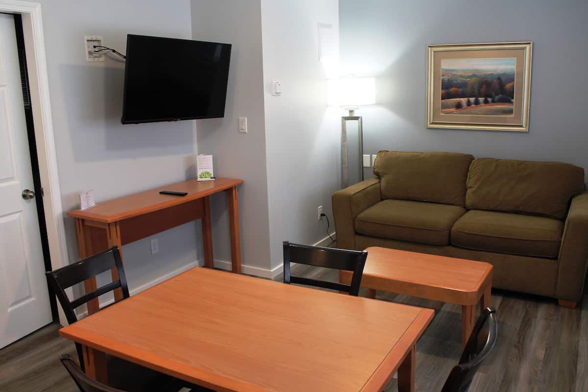 A living room with a brown sofa, a wooden dining table with chairs, a flat-screen TV mounted on the wall, and a framed landscape painting on the wall.
