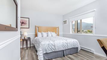 Un dormitorio con cama, mesita de noche con lámpara, ventana con vistas y un cuadro en la pared.