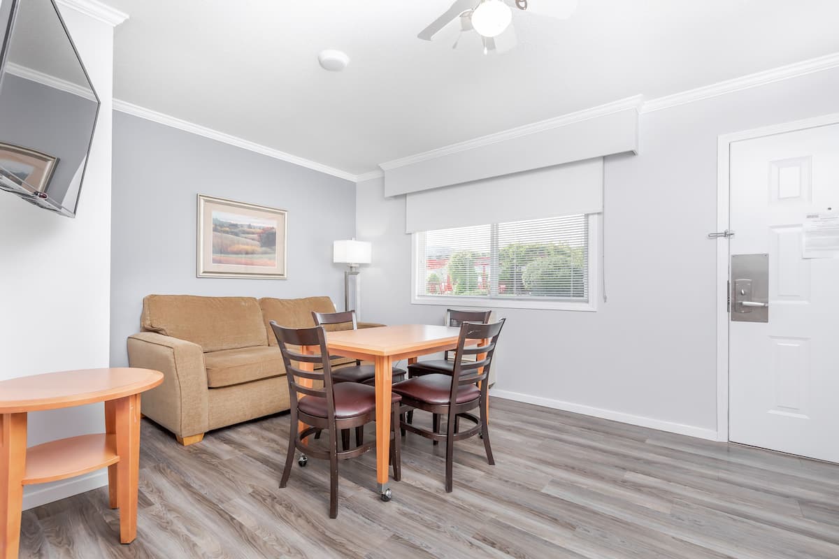 A room with a wooden dining table, four chairs, a sofa, a TV mounted on the wall, a window with blinds, and a door with a lock.