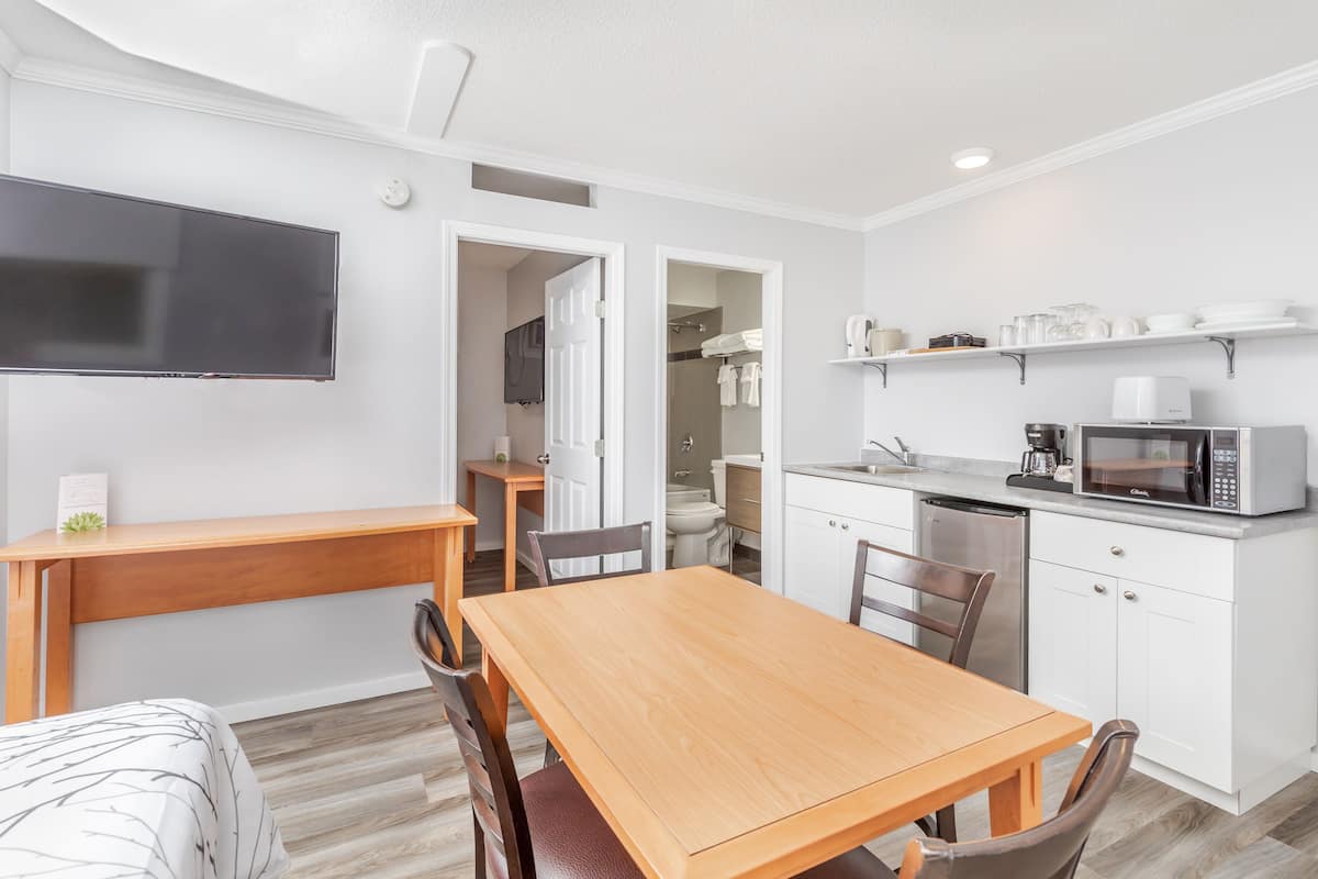 A modern kitchen with white cabinets, a microwave, and a coffee maker. There is a wooden dining table and chairs, and a TV mounted on the wall.