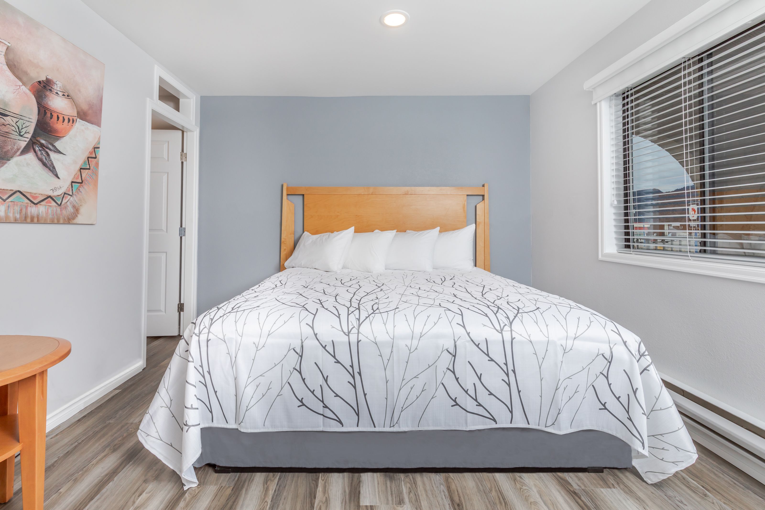 A bedroom with a wooden headboard, a bed with a white and gray tree-patterned bedspread, a wooden floor, a round wooden table, and a painting on the wall.