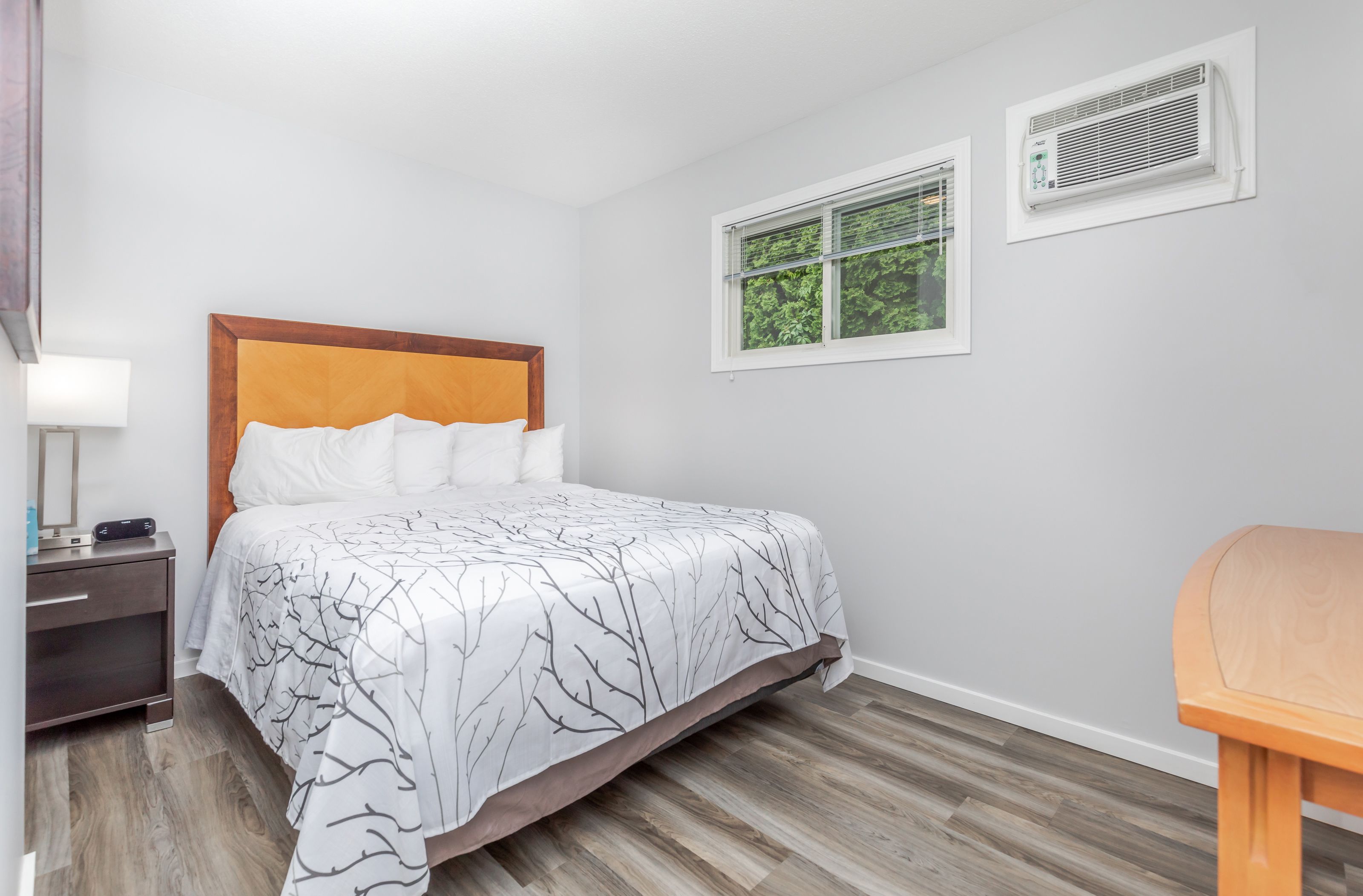 Una cama bien hecha con colcha de diseño blanco y negro en forma de árbol, un cabecero de madera y una mesita de noche con una lámpara y un pequeño dispositivo electrónico.