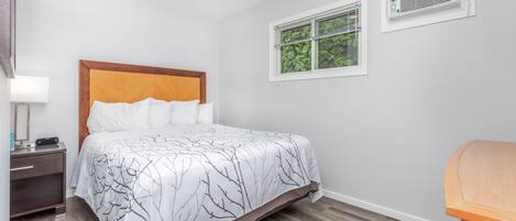 Una cama bien hecha con colcha de diseño blanco y negro en forma de árbol, un cabecero de madera y una mesita de noche con una lámpara y un pequeño dispositivo electrónico.