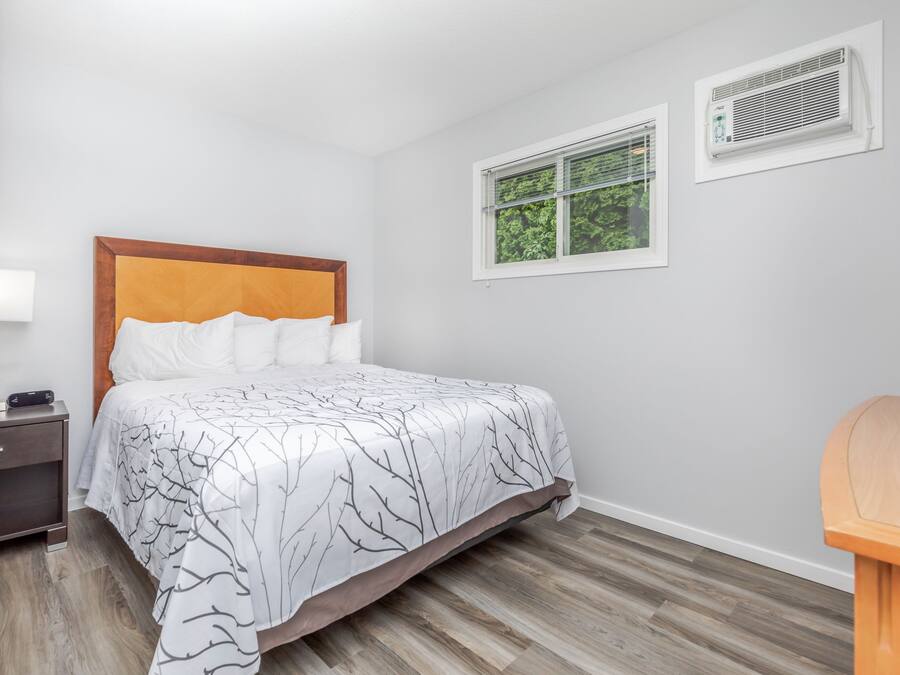 Un lit bien fait, recouvert d’une couvre-lit blanche et noire à motifs d’arbres, avec une tête de lit en bois et une table de chevet sur laquelle se trouvent une lampe et un petit appareil électronique.