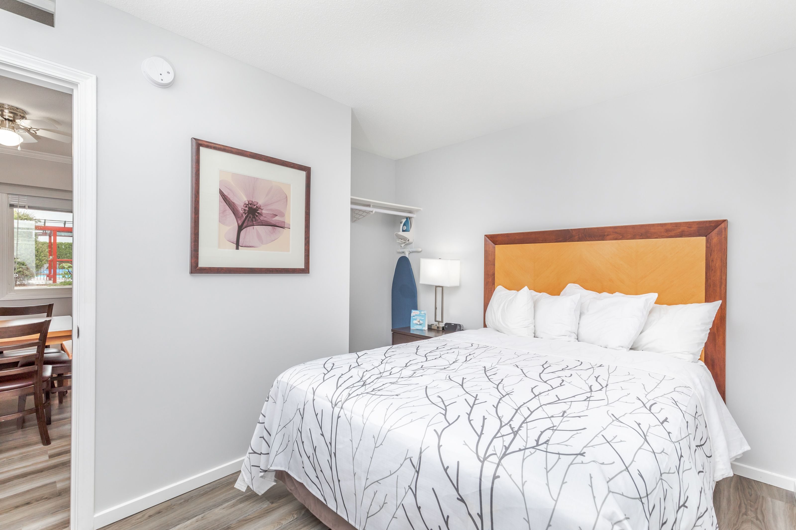 Una habitación de hotel con cama, mesita de noche, lámpara, un cuadro en la pared y una zona de comedor visible a través de una puerta abierta.
