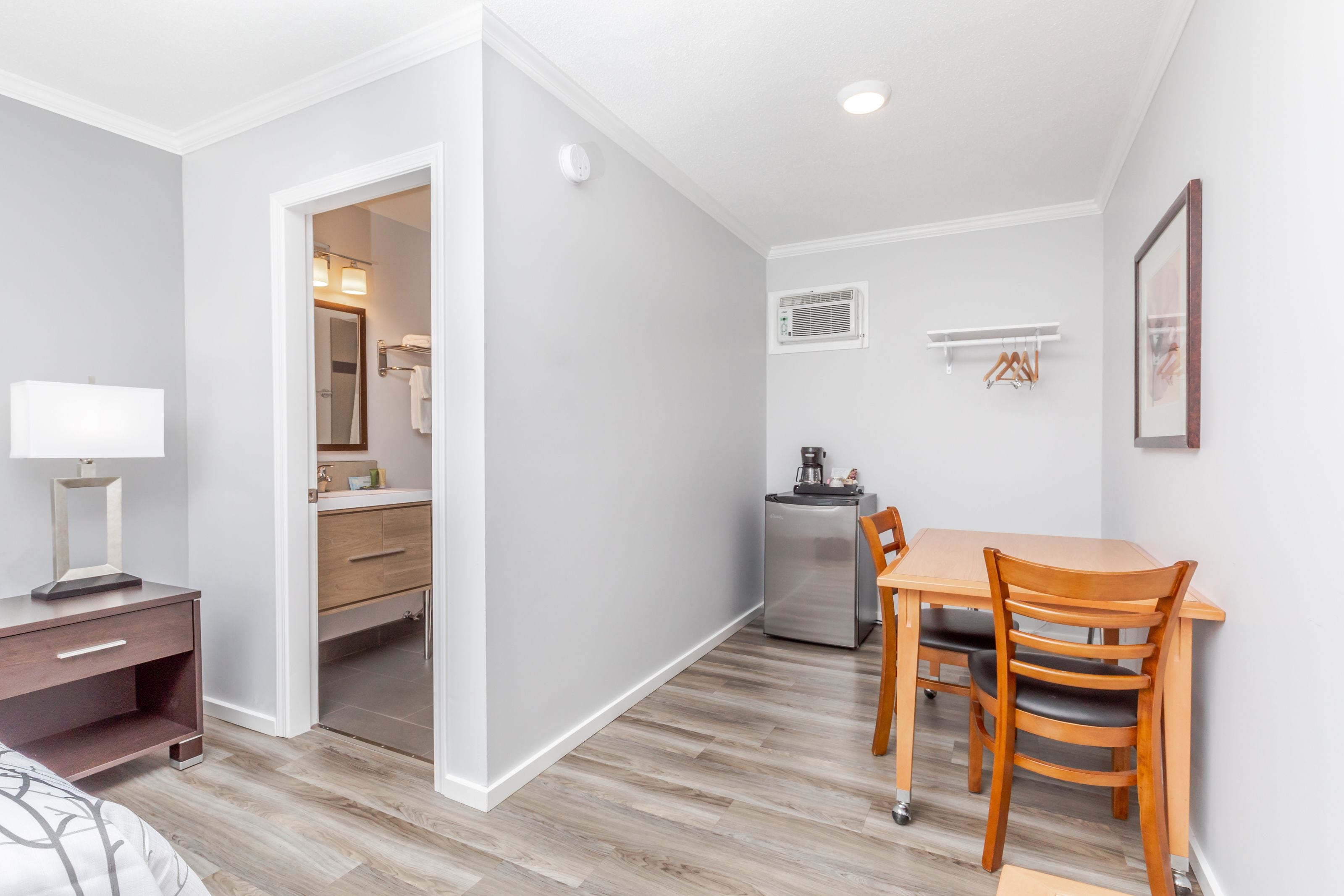 A compact room with a wooden dining table, chairs, a small kitchenette, a mirror, and a bedside table with a lamp.