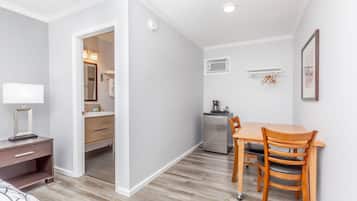 A compact room with a wooden dining table, chairs, a small kitchenette, a mirror, and a bedside table with a lamp.
