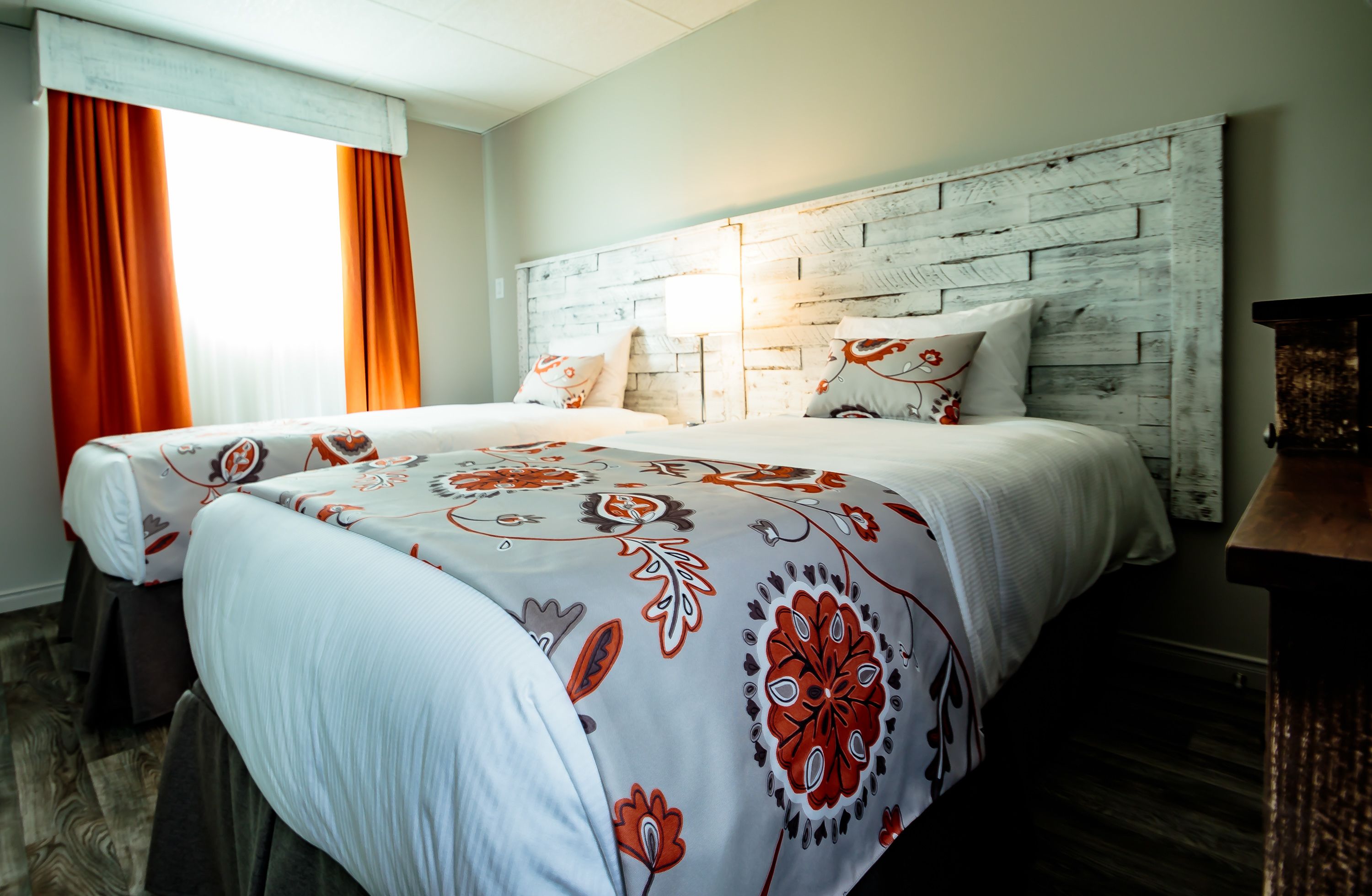 A hotel room with a rustic wooden headboard, two beds with floral-patterned bedding, and a large window with orange curtains.