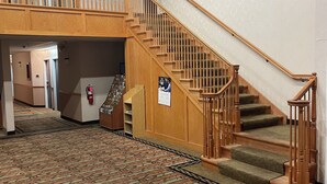 A hotel lobby with a patterned carpet, a wooden staircase with a glass railing, and a fire extinguisher on the wall.