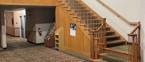 A hotel lobby with a patterned carpet, a wooden staircase with a glass railing, and a fire extinguisher on the wall.