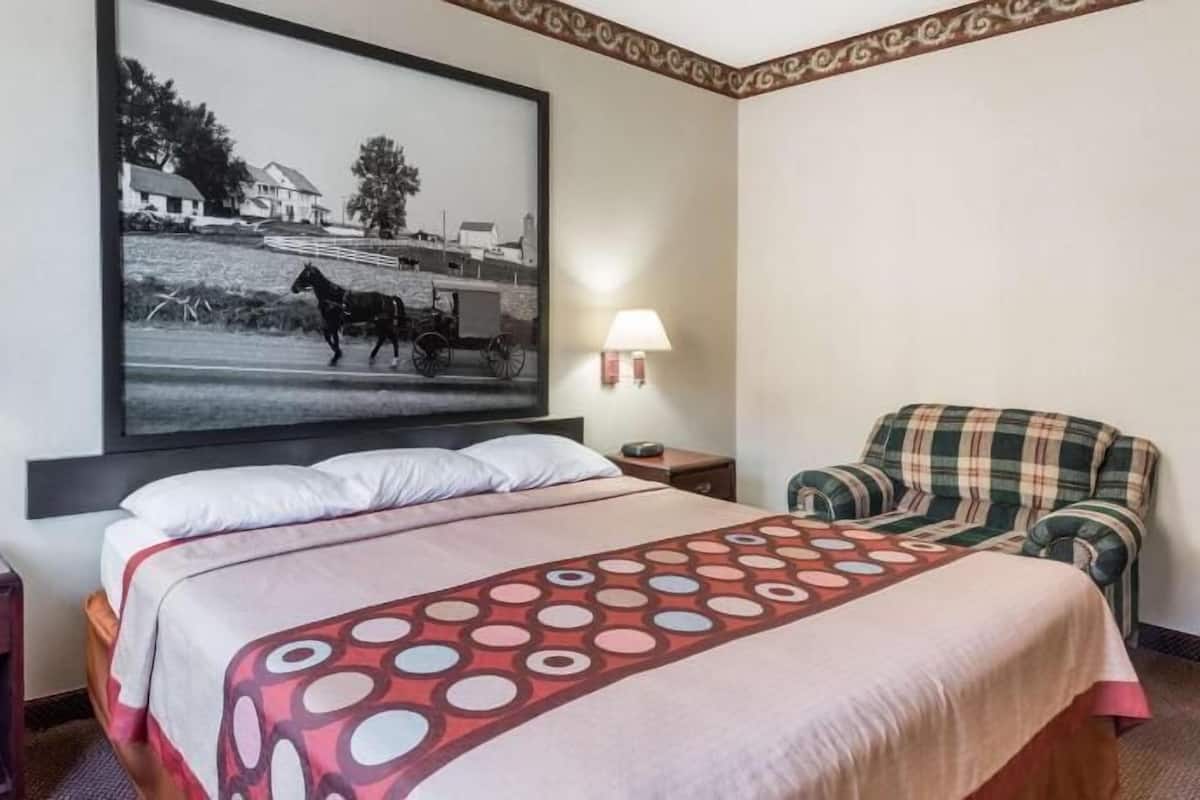 A hotel room with a bed, two bedside tables, a chair, a large framed picture on the wall, and two wall-mounted lamps.
