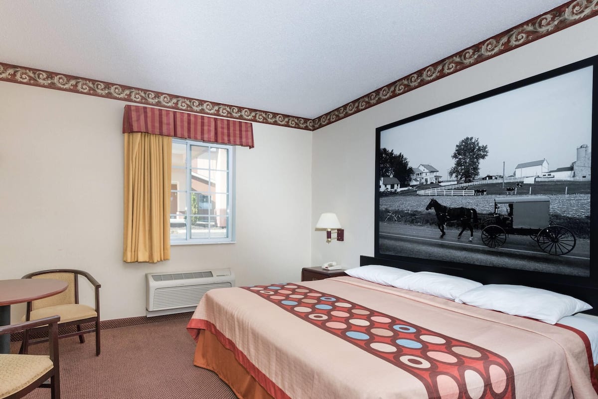 A hotel room with a bed, a chair, a table, a window with curtains, a wall-mounted lamp, and a large picture of a horse-drawn carriage.