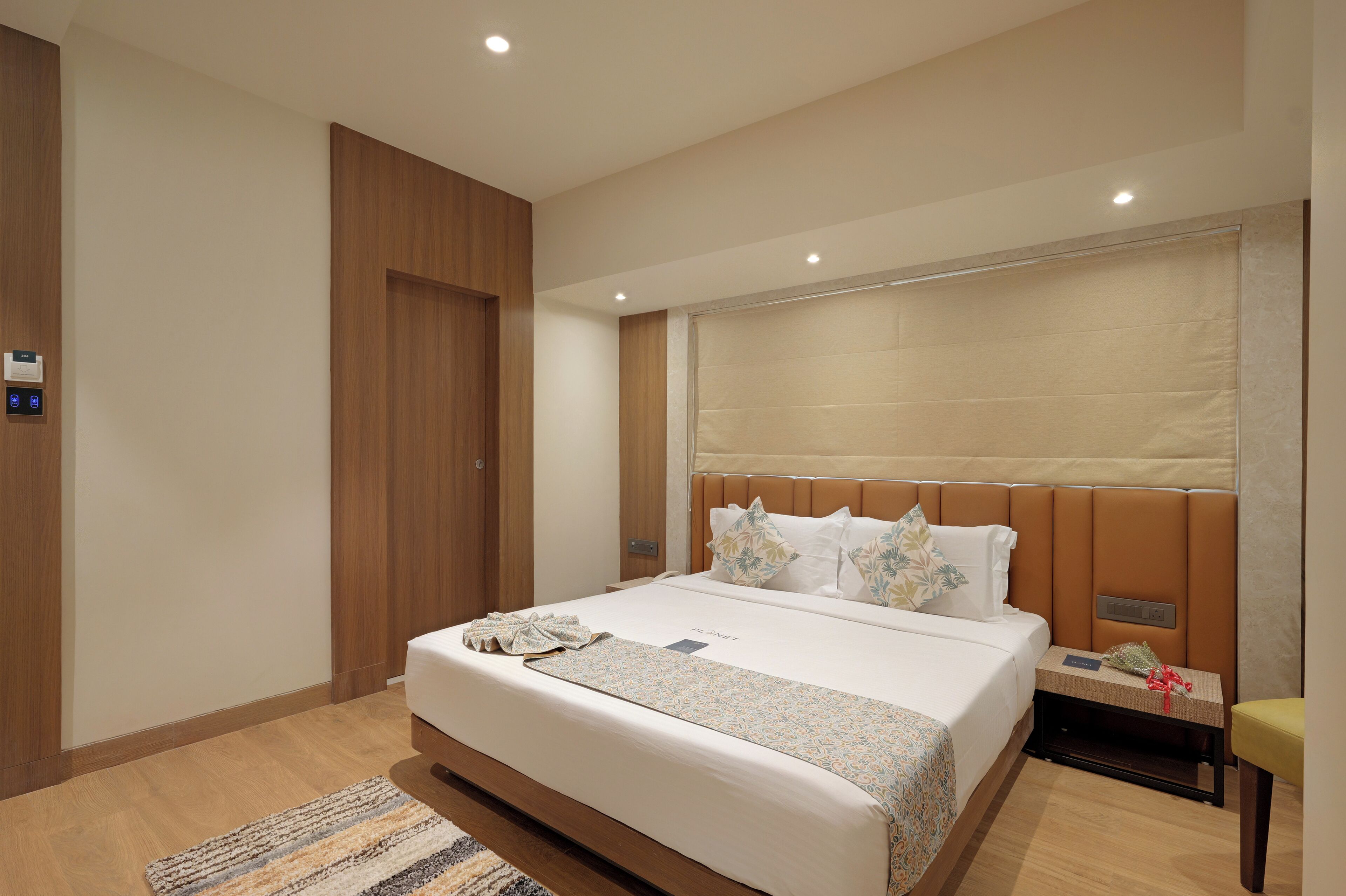 A modern hotel room with a large bed, a bedside table, a nightstand, a wooden wardrobe, and a ceiling light.