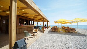 Una terrassa de fusta amb seients exteriors i para-sols, amb vista a una platja de petits còdols.
