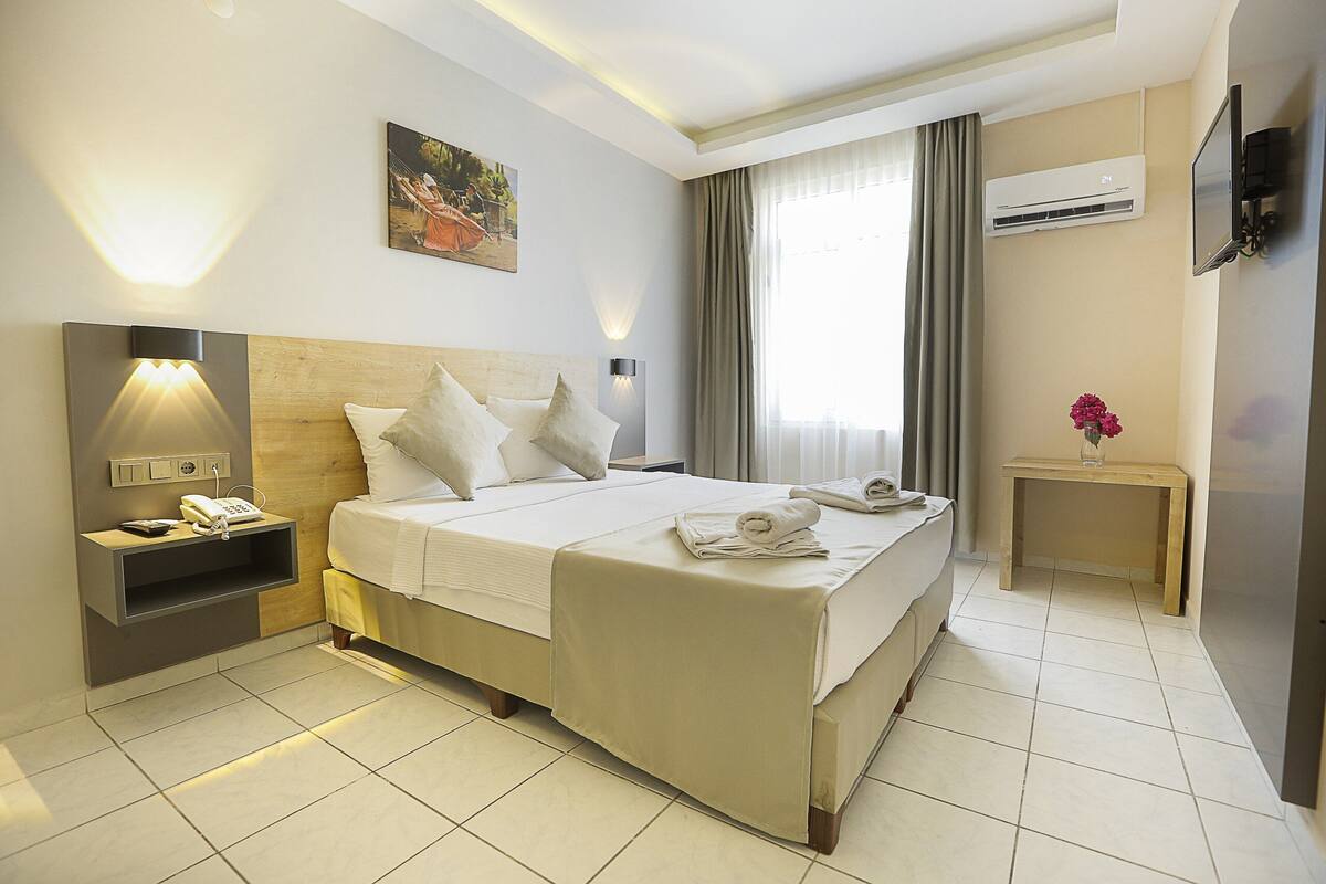 Une chambre d’hôtel comprenant un lit, une table de chevet, une lampe, un appareil de climatisation et une petite table avec un vase de fleurs.