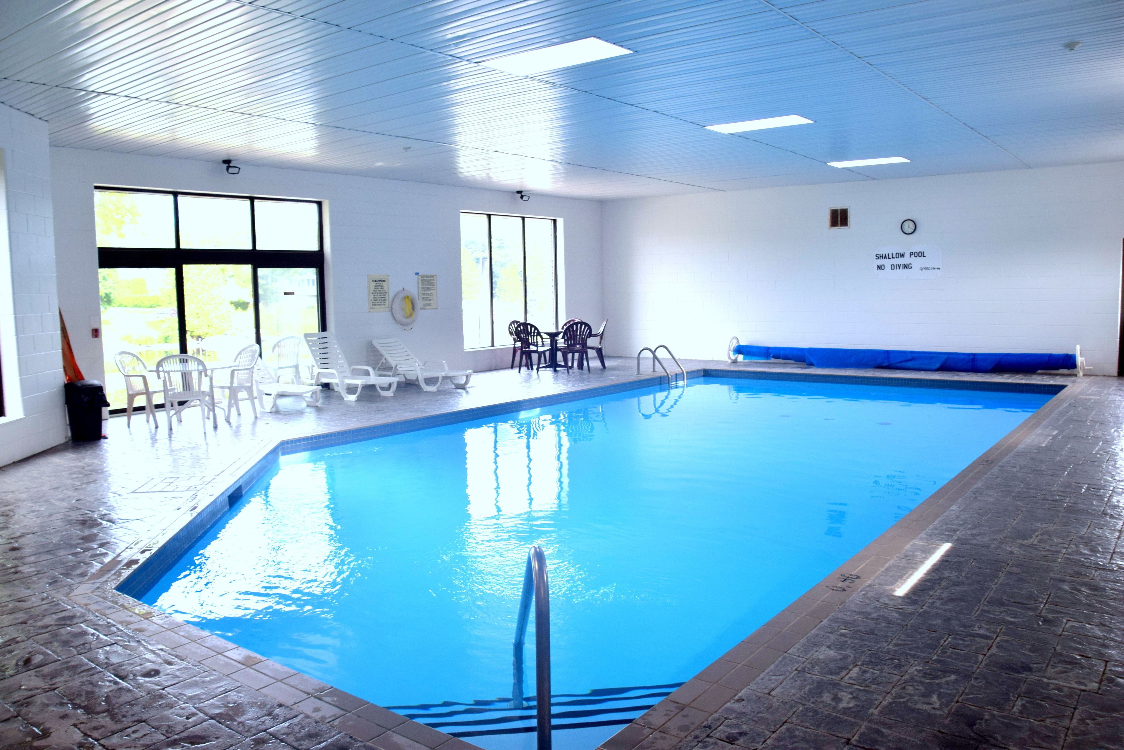 Una piscina interior con agua azul cristalina, un cartel de advertencia para aguas poco profundas y varias sillas reclinables blancas alrededor de la piscina.