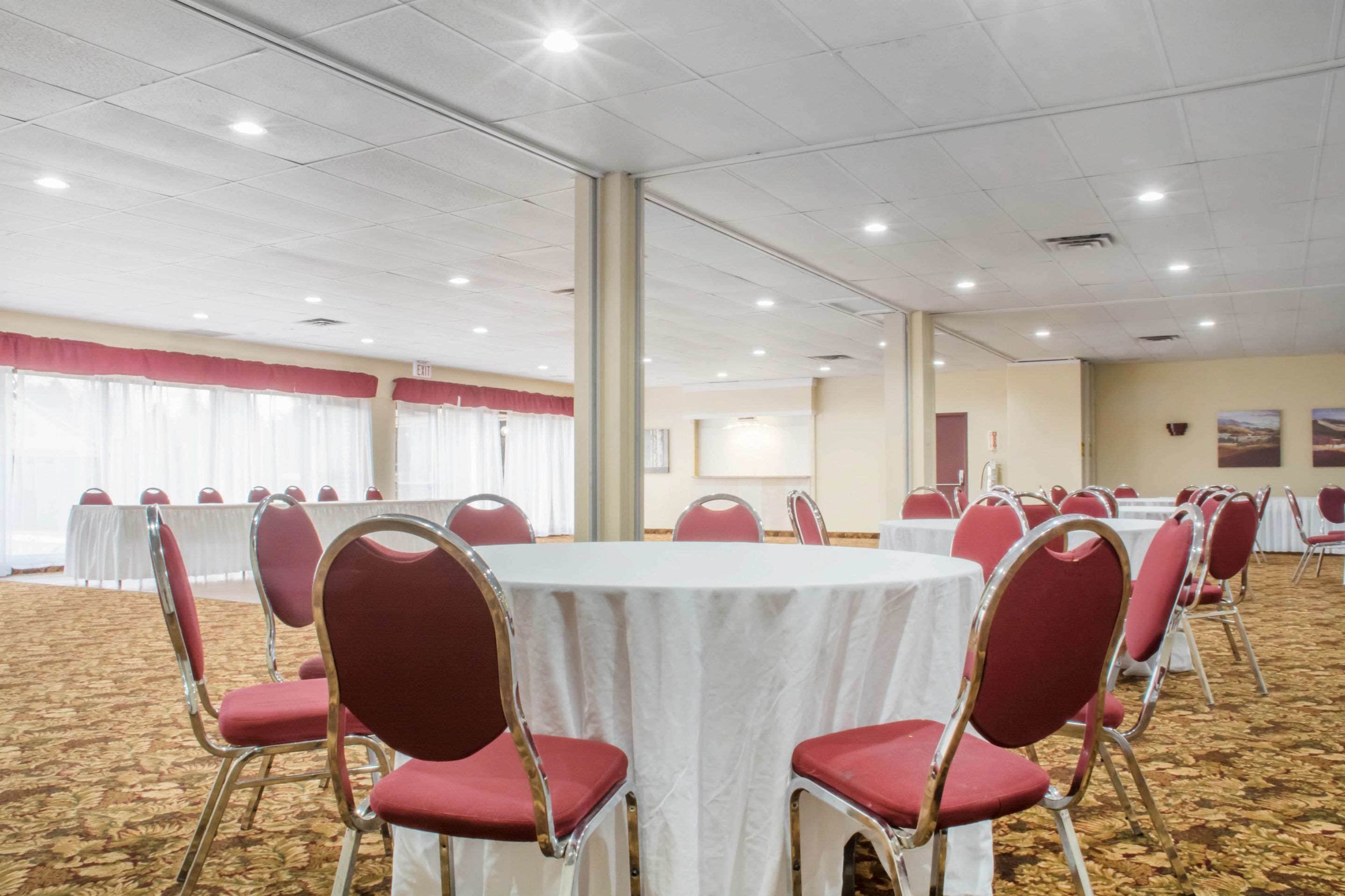 Un salón de conferencias con mesas redondas y sillas rojas, piso alfombrado y cortinas blancas.