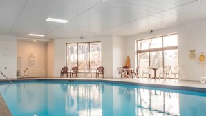 An indoor swimming pool with clear water, surrounded by white chairs and tables, large windows, and ceiling lights.