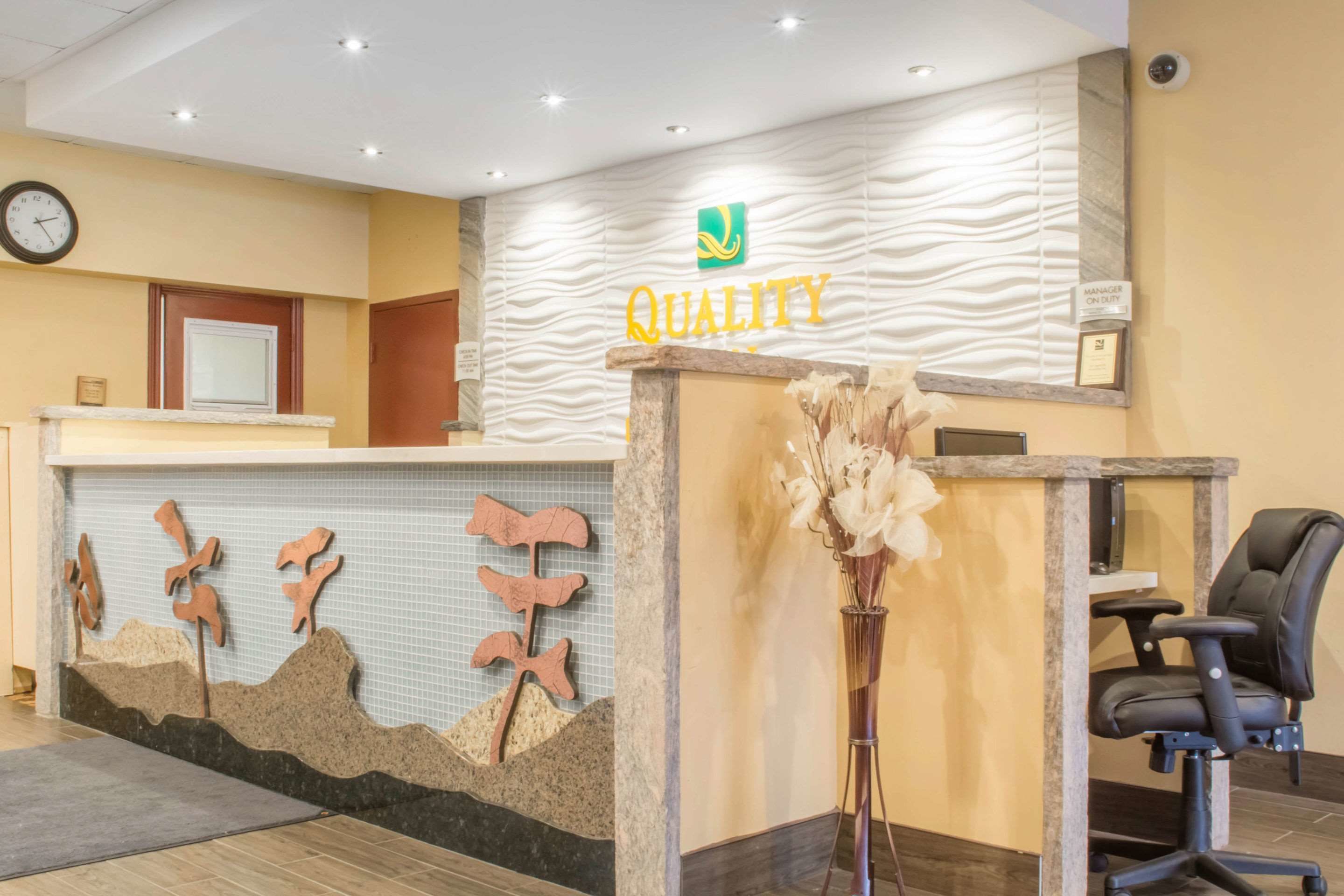 Una recepción con un letrero que dice 'QUALITY', un reloj y una pared decorativa con formas abstractas.
