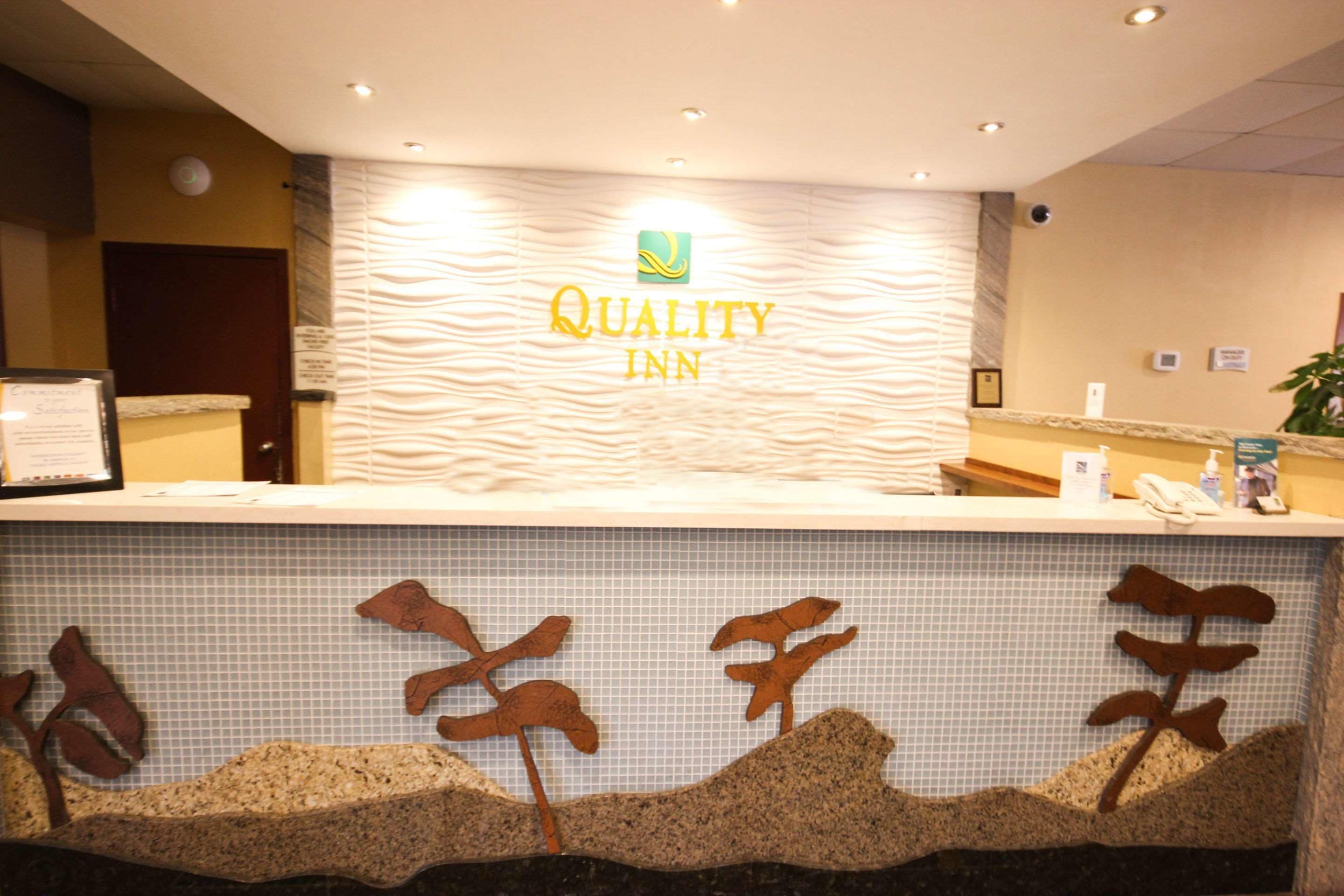 Un taulell de recepció amb el rètol 'Quality Inn', una zona de recepció amb una paret de rajoles i una planta a la dreta.