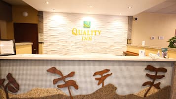 Un comptoir d’accueil avec une enseigne « Quality Inn », un espace de réception avec un mur carrelé et une plante sur la droite.