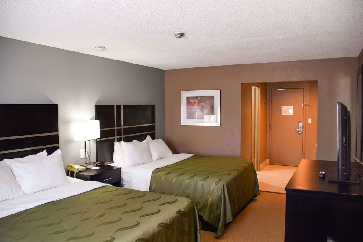A hotel room with two beds, a desk, a television, and a door leading to a hallway.