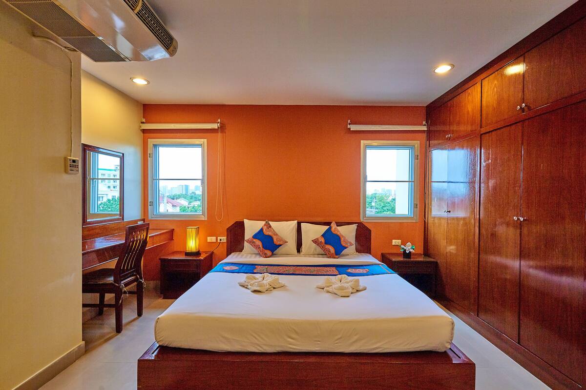 Habitación de hotel con una cama grande, dos mesitas de noche, un escritorio y una silla. Hay dos ventanas con vistas a la ciudad.