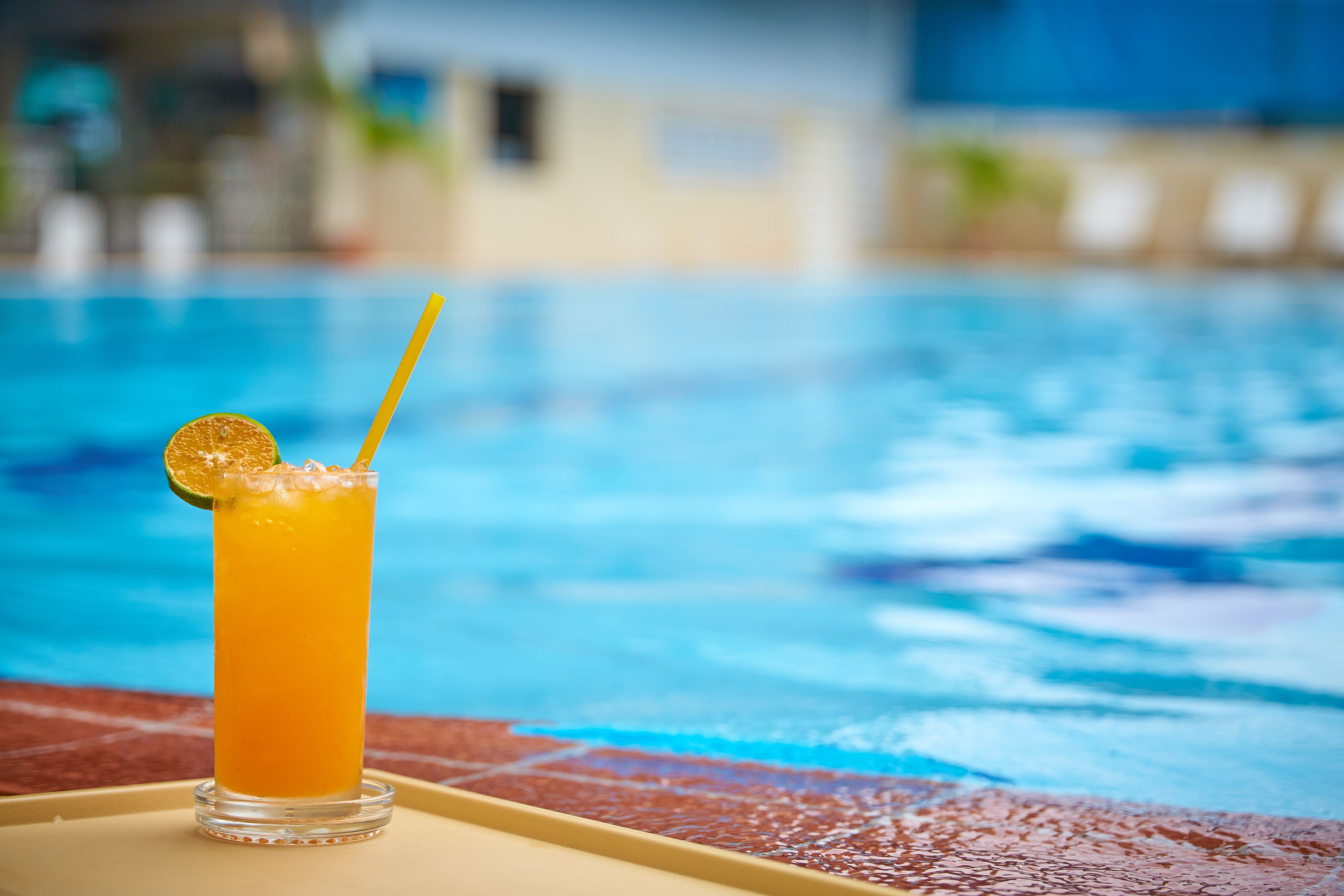 A glass of orange juice with a straw and a slice of lime on a tray by a pool.