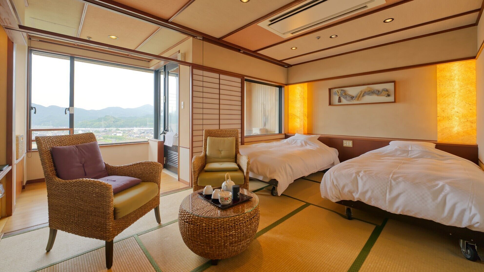A hotel room with two beds, a wicker chair, a small round table with tea set, and a view of mountains through large windows.
