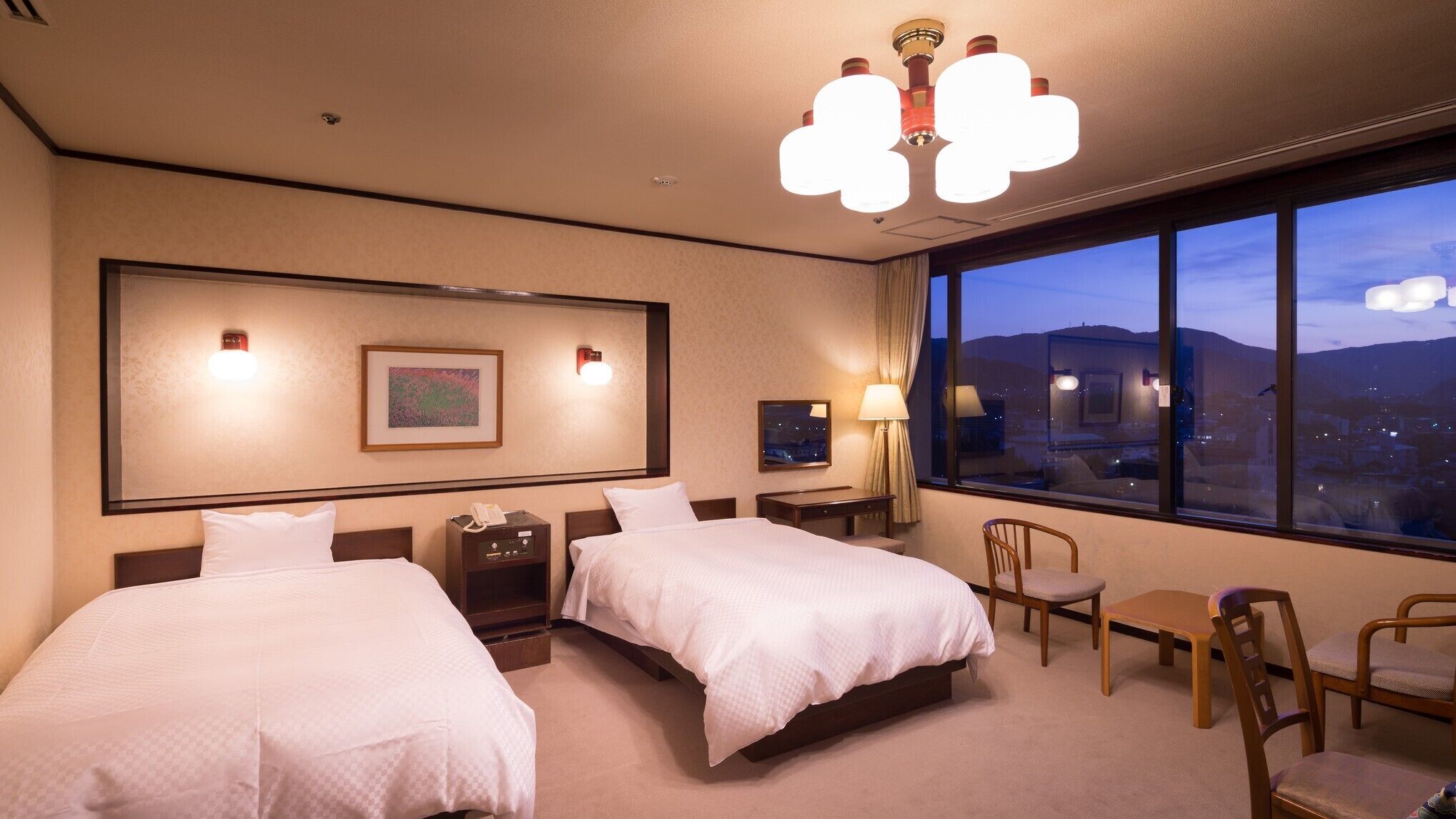 A hotel room with two beds, a desk, chairs, a large window with a city view, and a chandelier.