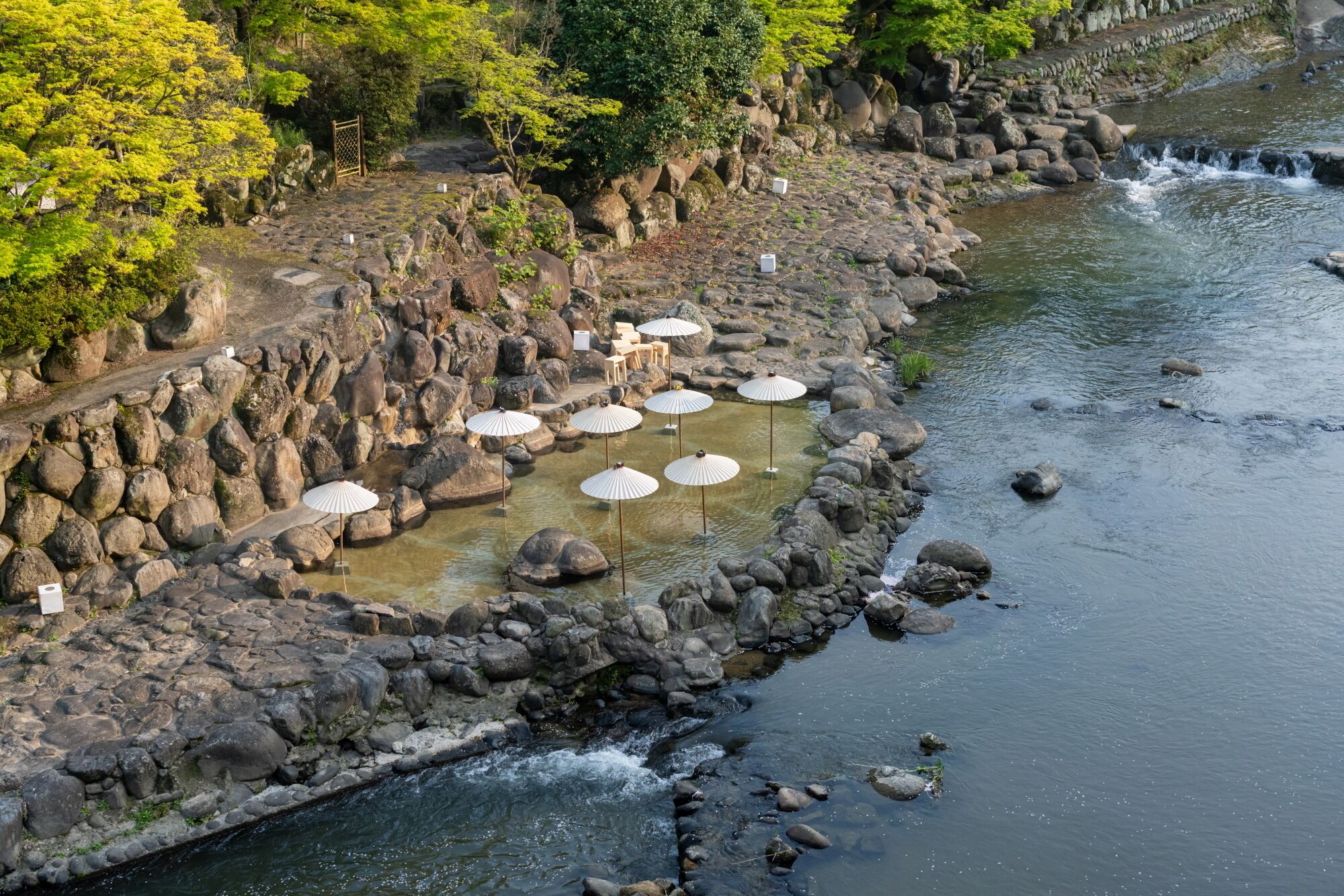 Un paseo ribereño con rocas, una pequeña cascada y varias sombrillas blancas.