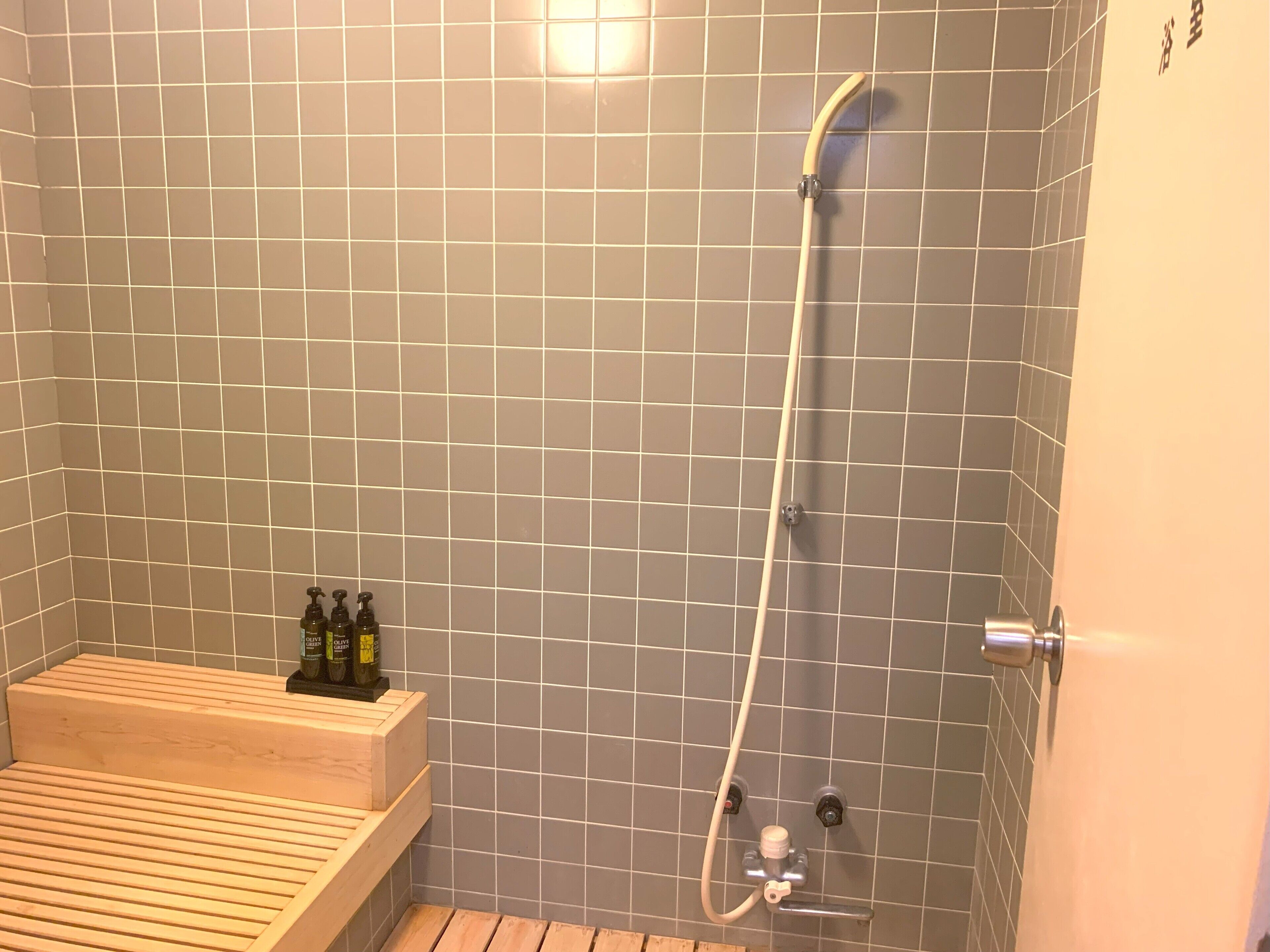 A small, tiled shower room with a wooden bench and a shelf holding bottles.