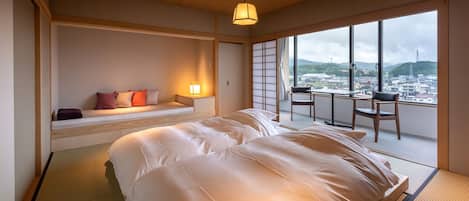Un dormitorio con una cama grande, una zona de estar con cojines, una mesa y vistas a un paisaje urbano a través de grandes ventanales.