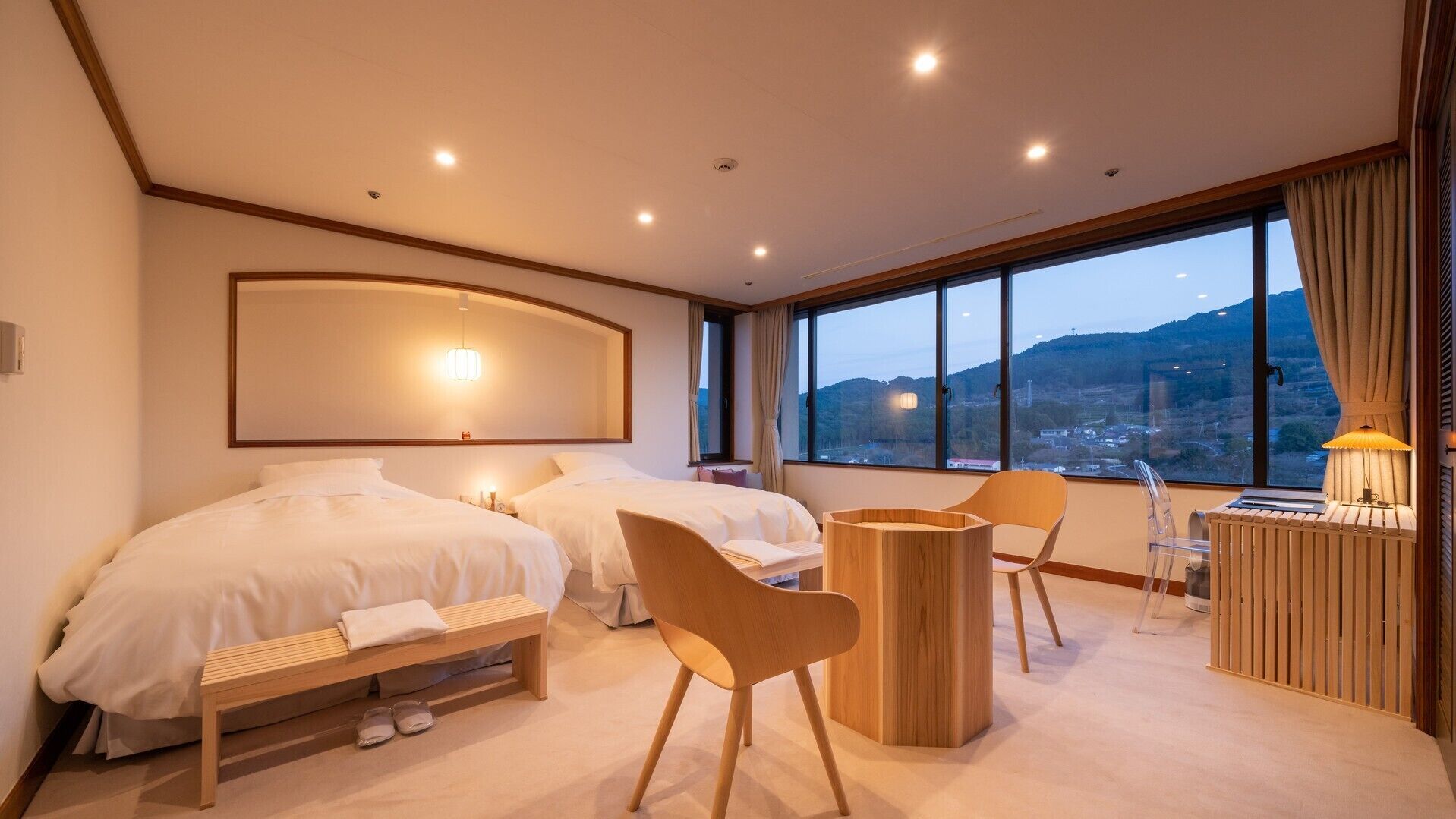 A hotel room with two beds, a desk, a chair, a bench, and a view of the mountains.