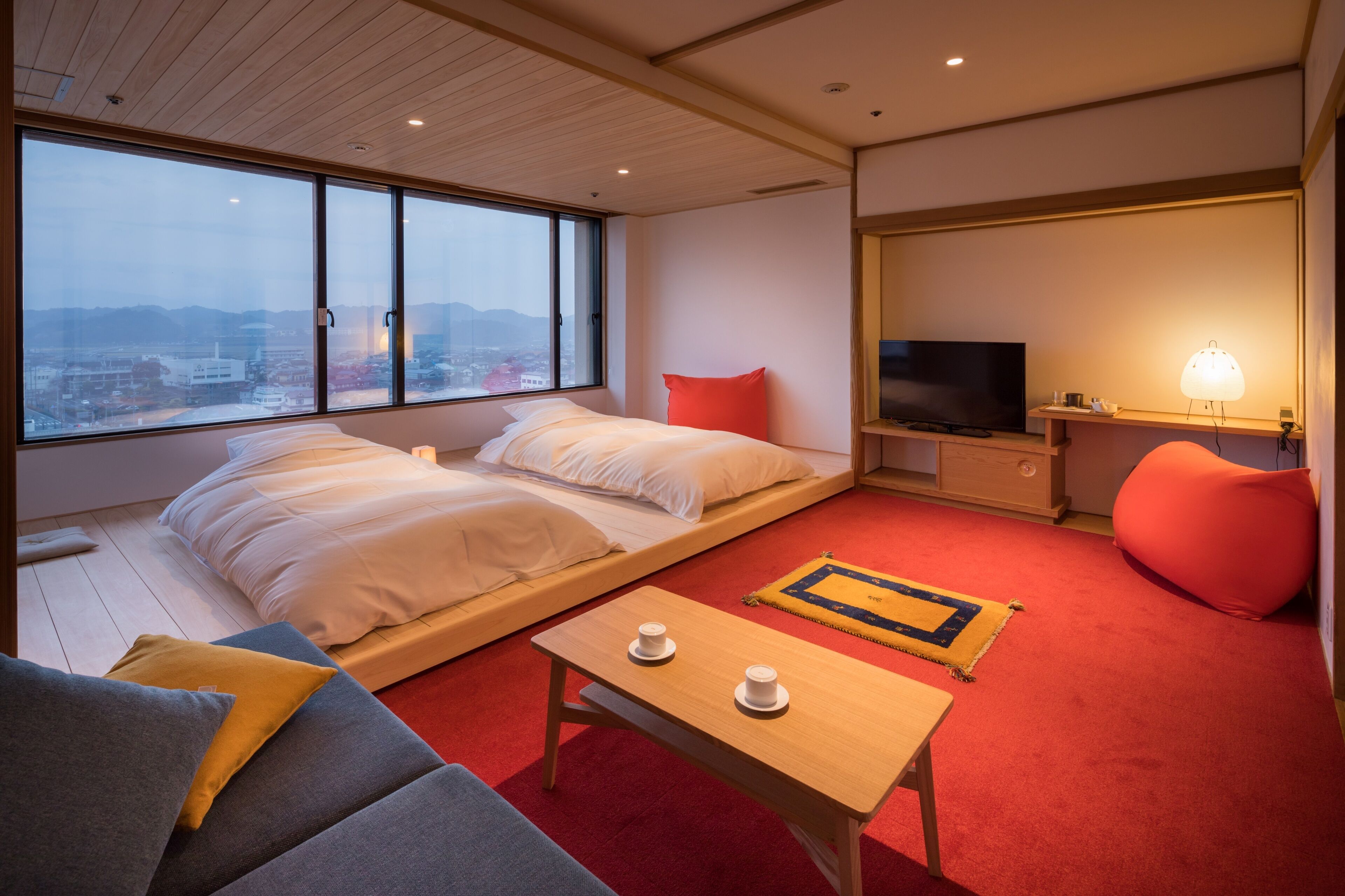 A modern hotel room with a large bed, a TV, a small table with cups, and a view of the city.