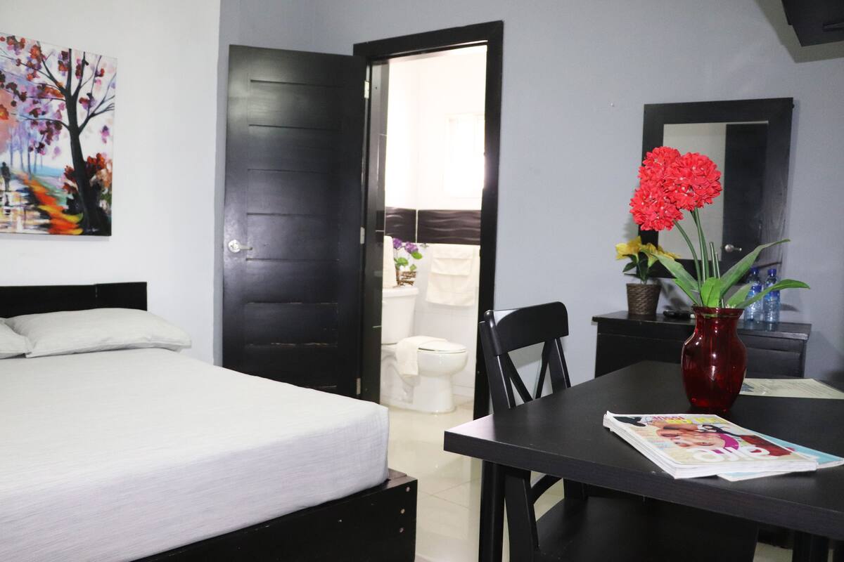 A bedroom with a bed, a table with a magazine, a red vase with flowers, and a painting on the wall.