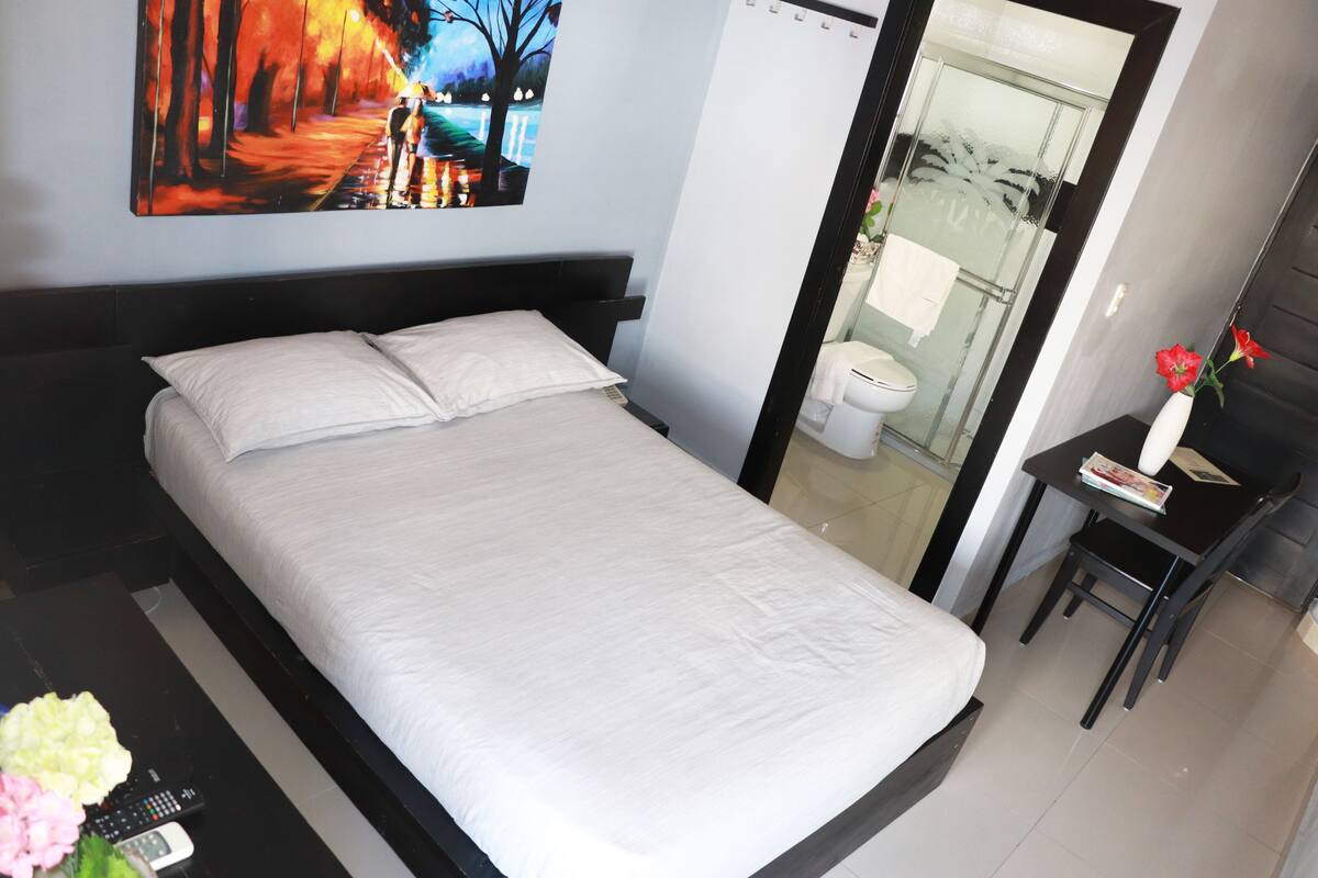 A hotel room with a bed, a painting on the wall, a bathroom visible through a glass door, and a small table with a vase of flowers.