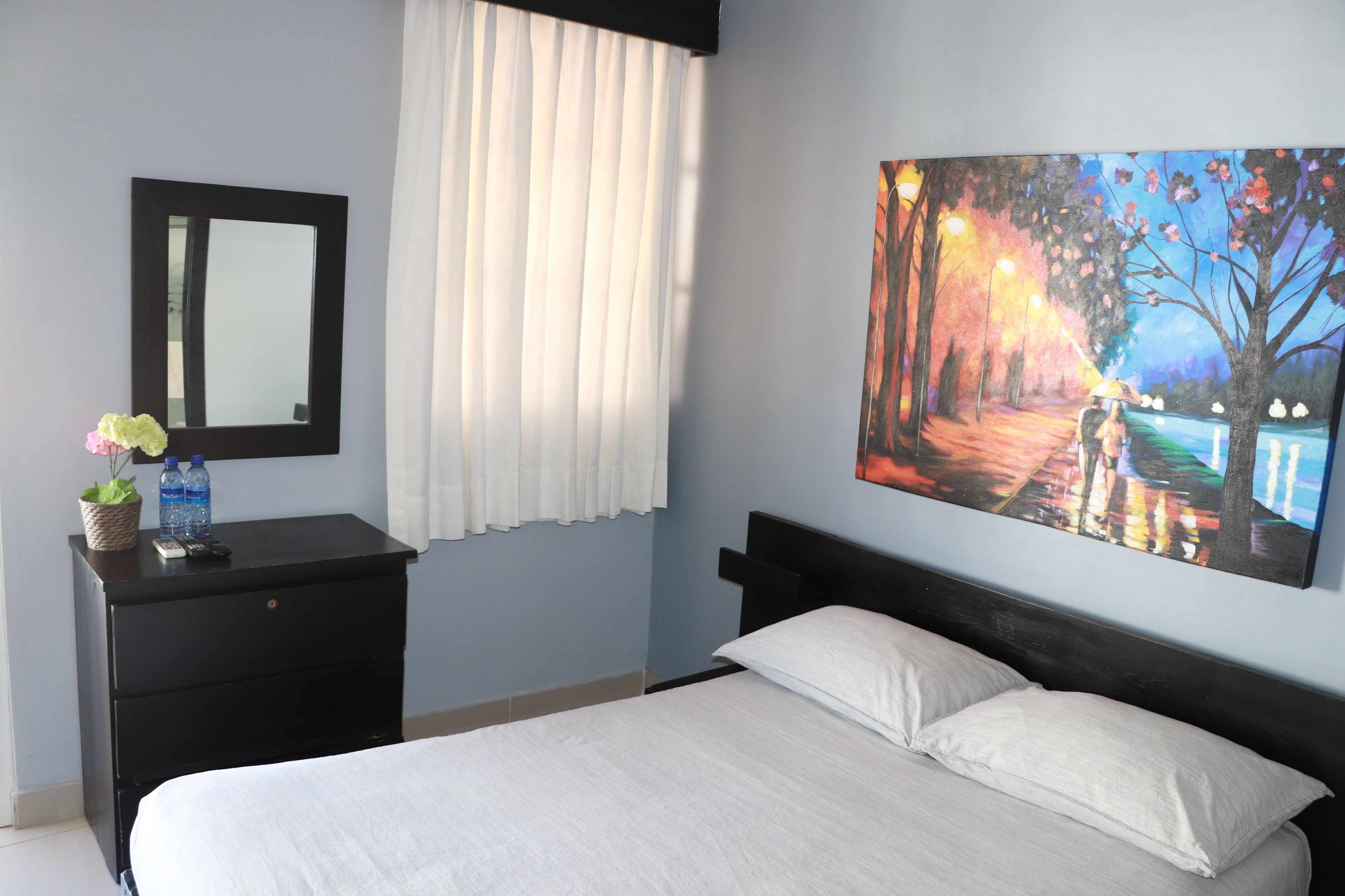 A bedroom with a bed, a nightstand, a mirror, a painting, and a window with curtains.