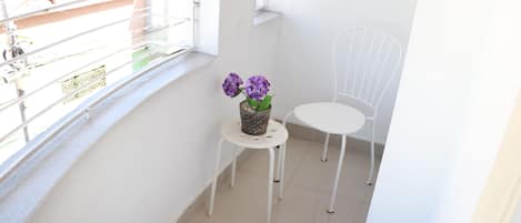 Un balcón con una silla blanca, una mesita con un cesto de flores y vistas a edificios al fondo.