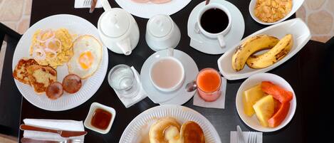 Una mesa de desayuno con platos de huevos, tocino, panqueques y tostadas. También hay tazas de café, un vaso de jugo y un tazón de cereal.