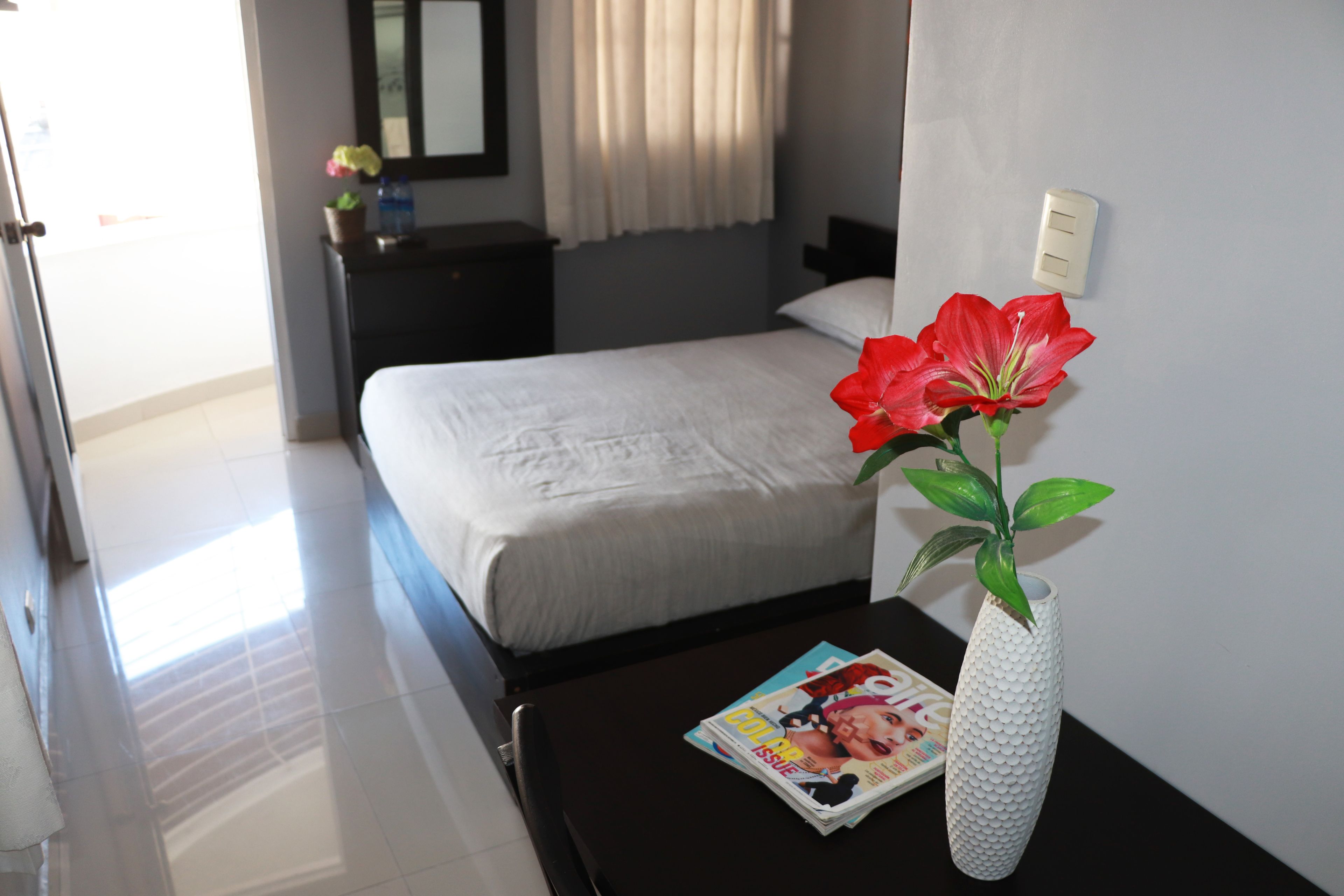 Une chambre d’hôtel avec un lit, une table de chevet, un magazine et un vase blanc contenant une fleur rouge.